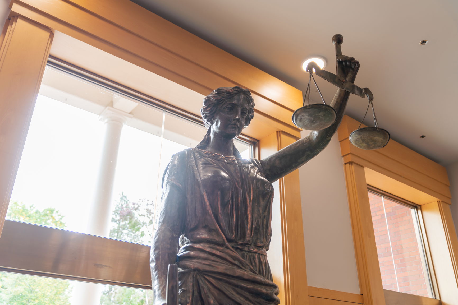 Bronze statue of Lady Justice holding balance scales in front of a large window.