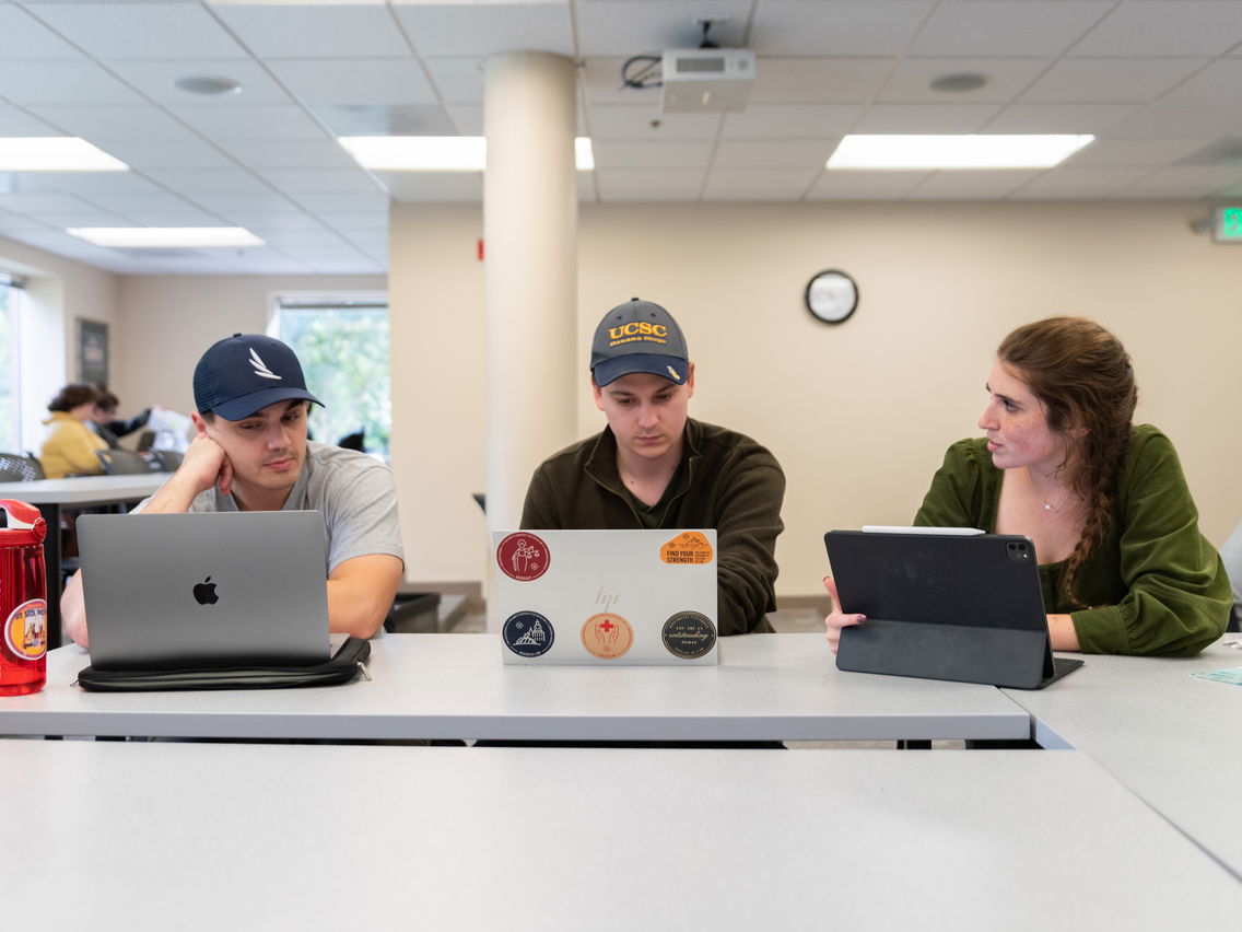 Three individuals sitting at a table, each using a laptop or tablet in a study area.