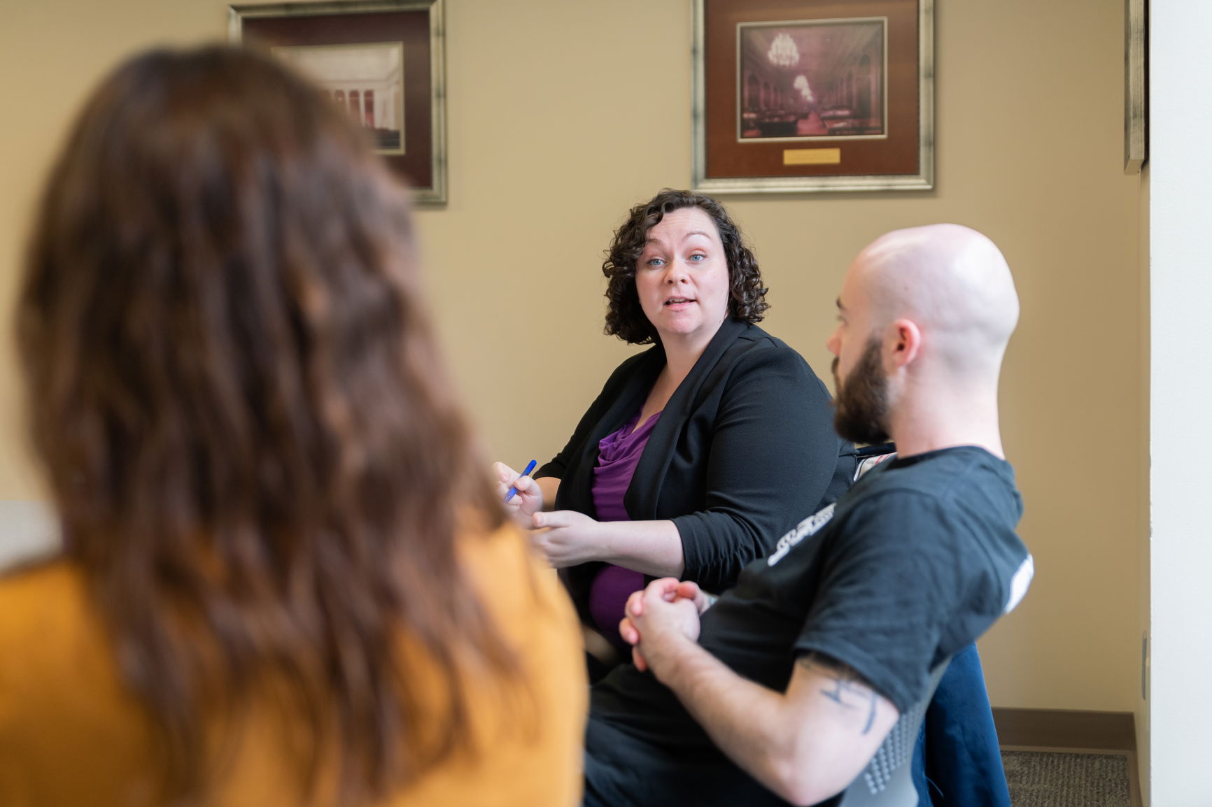 Three people seated in a discussion, focusing on a woman talking with a pen in hand.