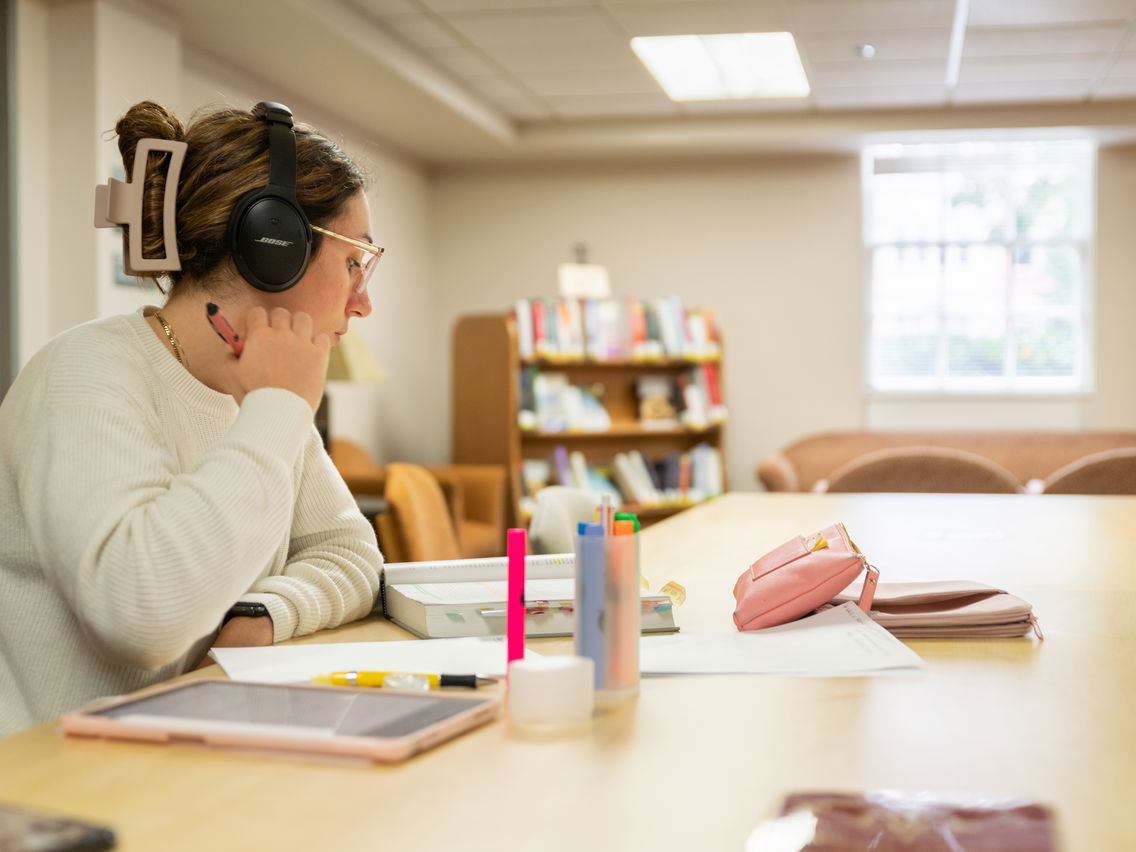 Person wearing headphones studying at a table with books and stationery in a library.