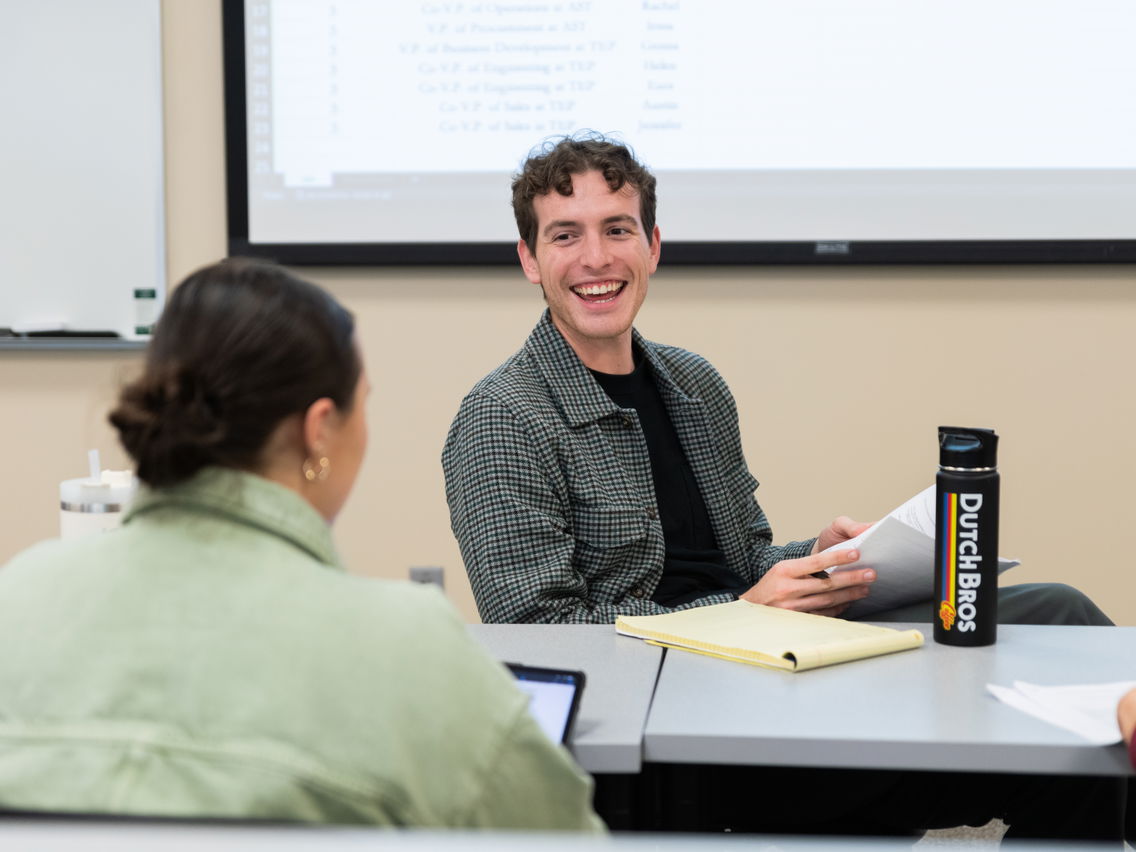 Man smiling and holding papers in a classroom, with a Dutch Bros water bottle and a notepad on the table.