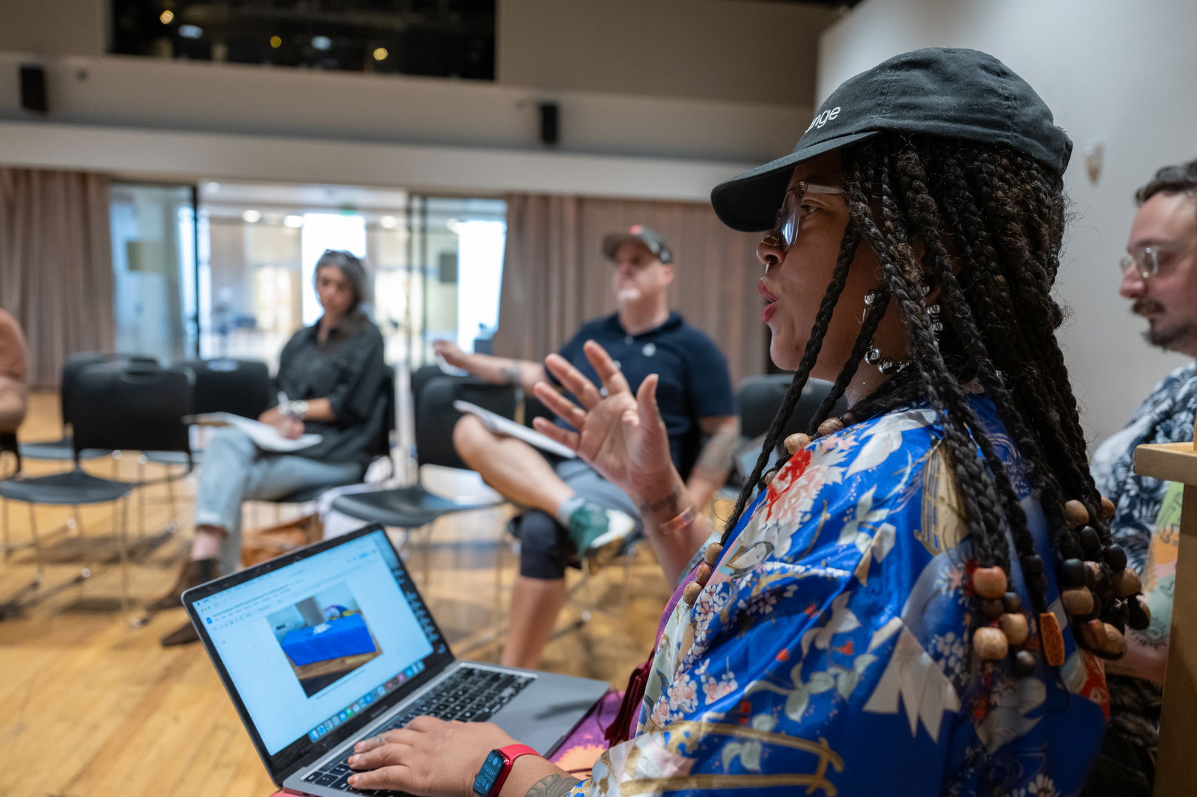 A person with a hat chatting to a classroom and a virtual meeting on their laptop using expressive hand gestures 