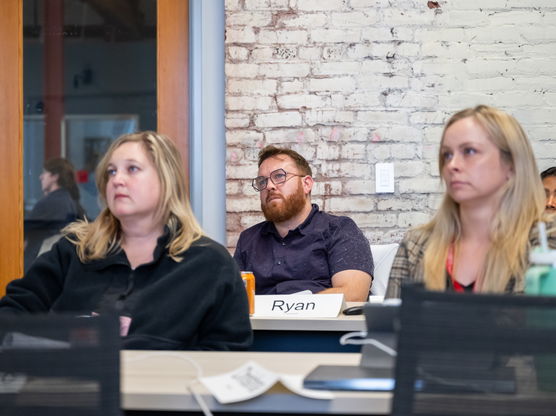 A classroom of people, focusing on two blonde students and a student with glasses