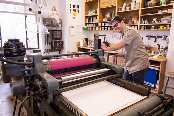 Man operating a vintage printing press in a well-lit printmaking studio, surrounded by jars and supplies on shelves.