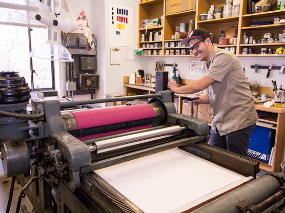 Man operating a vintage printing press in a well-lit printmaking studio, surrounded by jars and supplies on shelves.