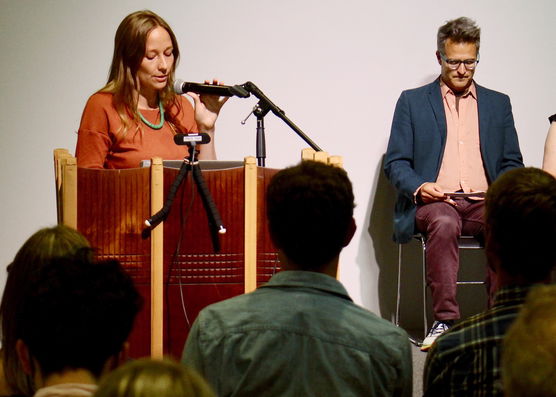 A guest reading with a faculty member sitting in the background. 