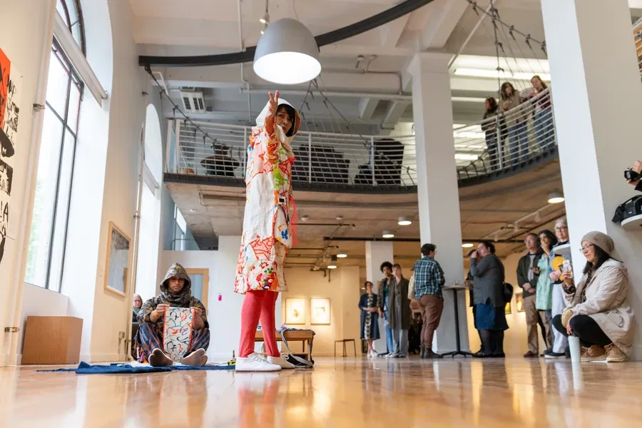 A performer gestures during an art event in a gallery, with an audience observing.