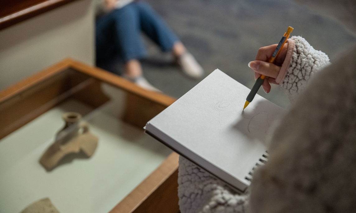 A closeup of a student's hand sketching something nearby.