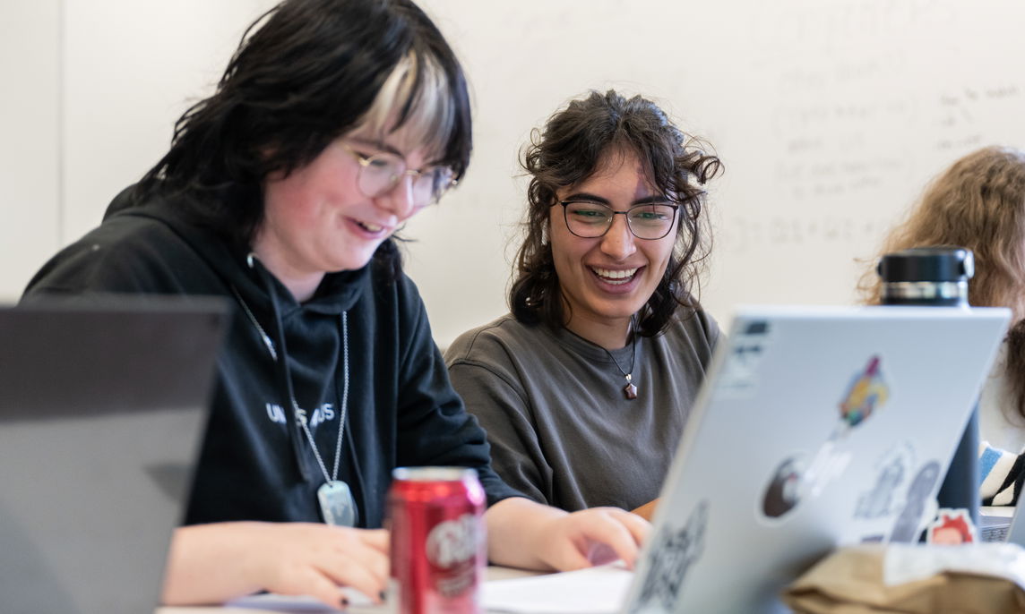 Two students laughing while looking at one laptop in a classroom