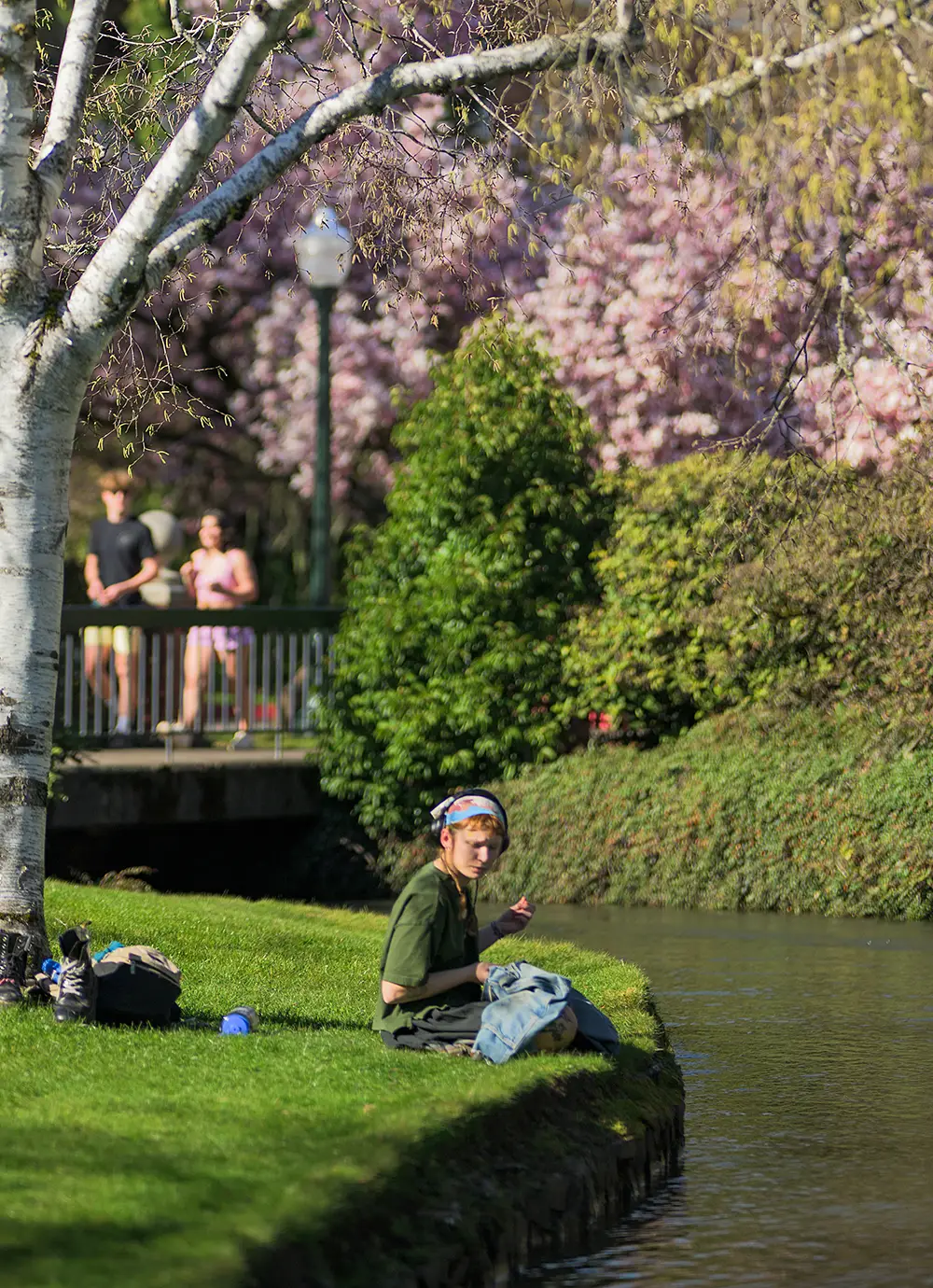 A Willamette University student sitting by the Mill Stream and two more students walking across the bridge in the background at Salem campus.