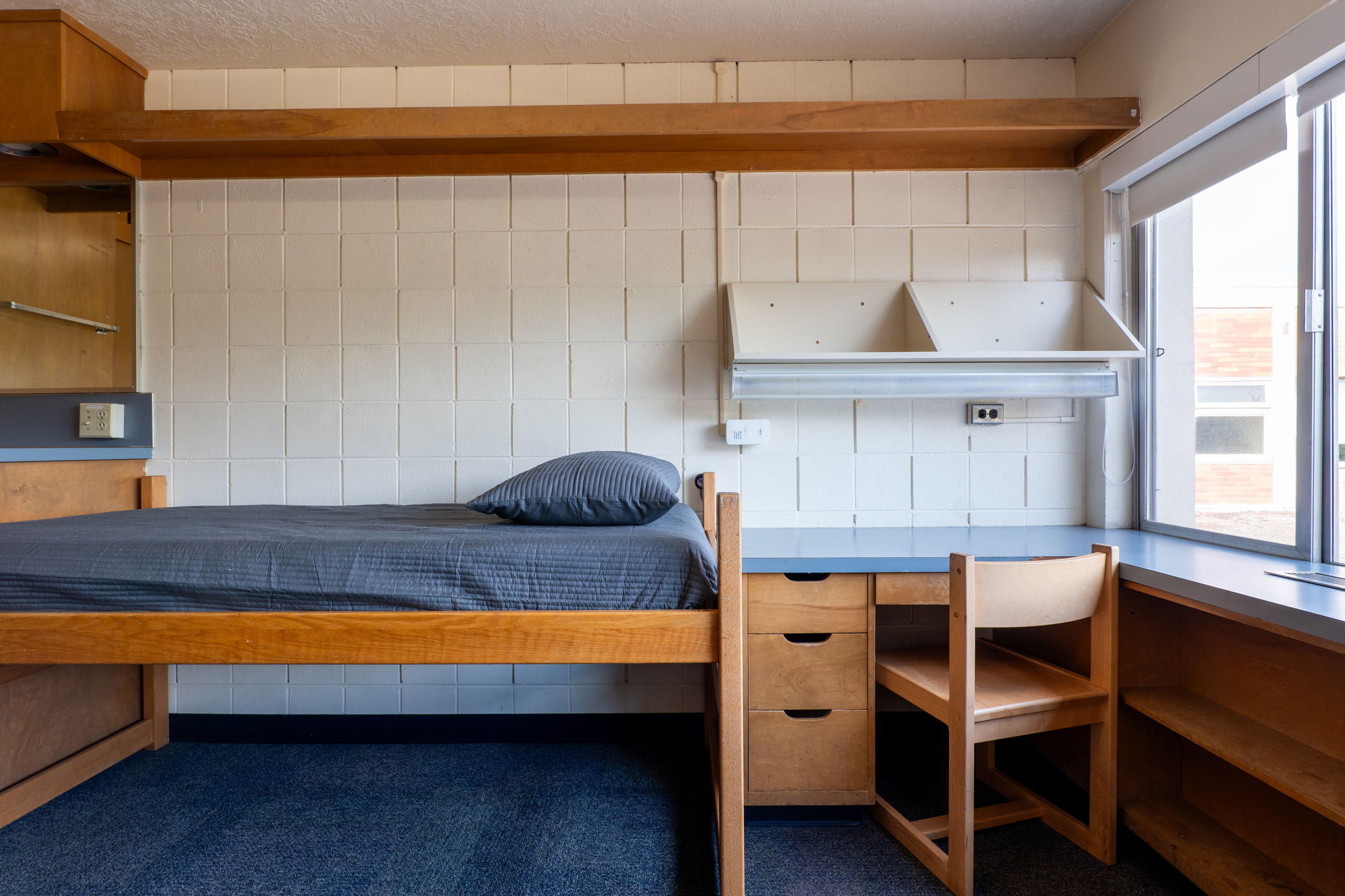 One side of a double room with a desk, bed, shelves, and chair