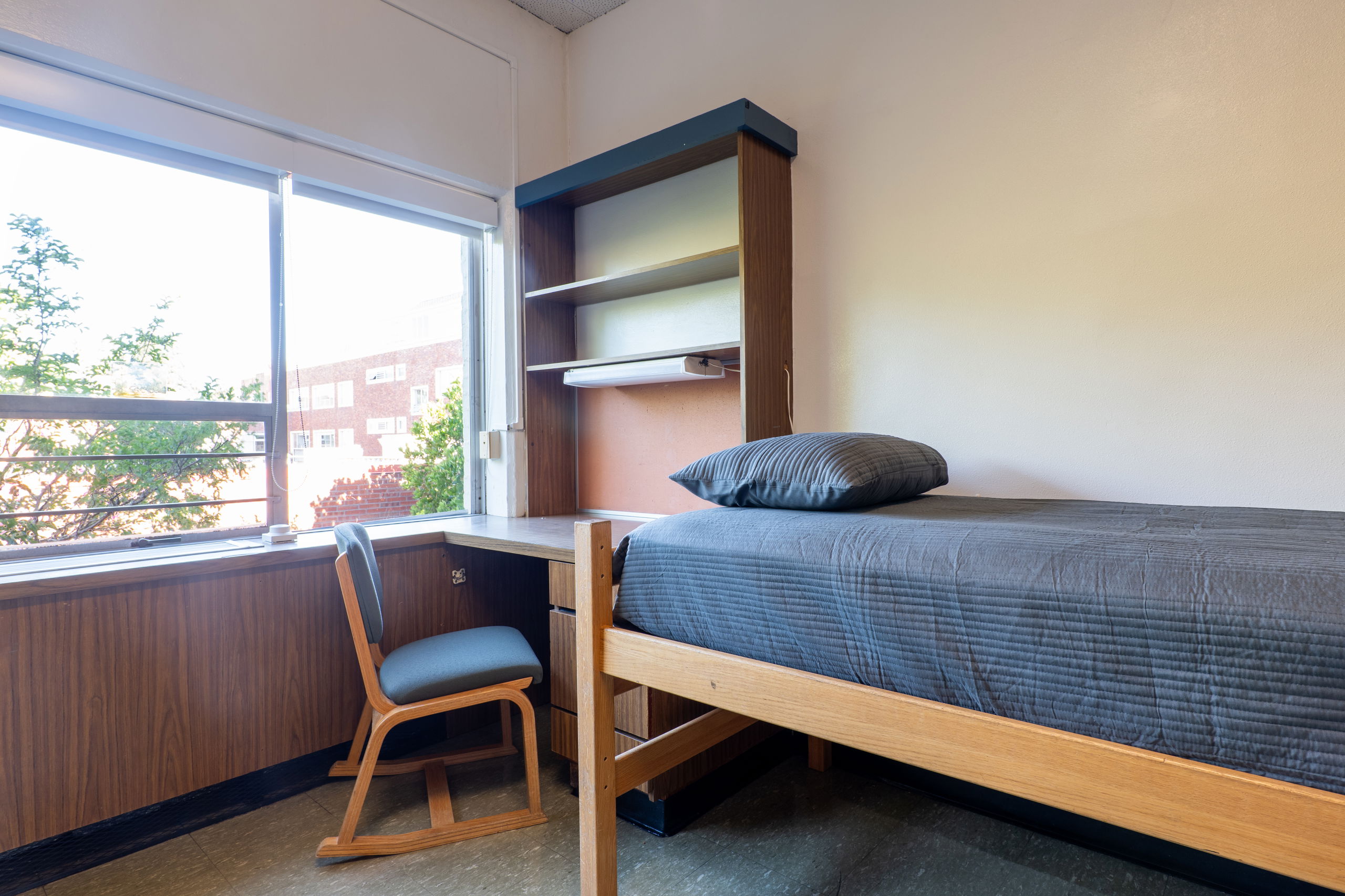 One side of a double room with a desk, bed, shelves, and chair