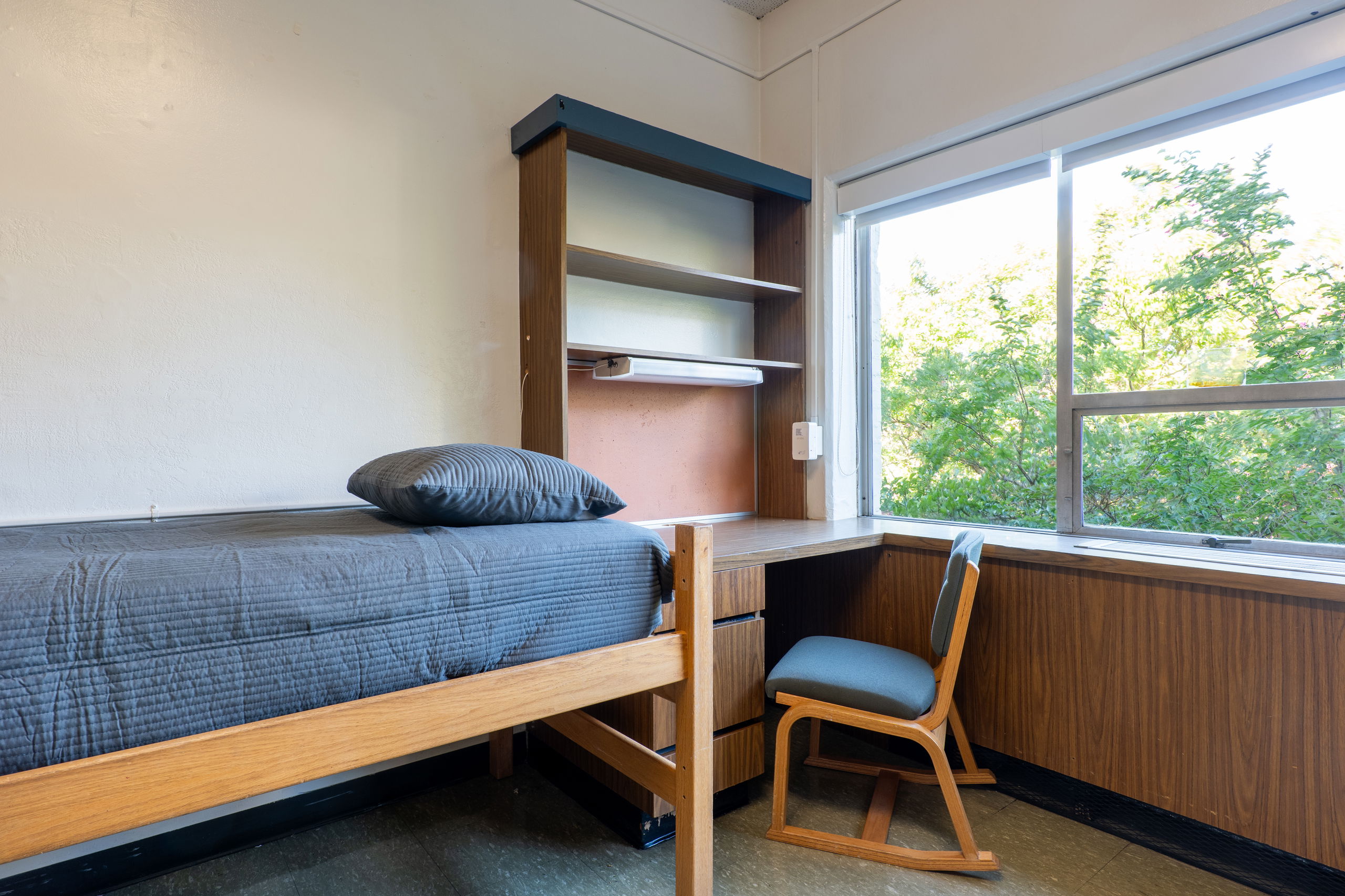 One side of a double room with a desk, bed, shelves, and chair