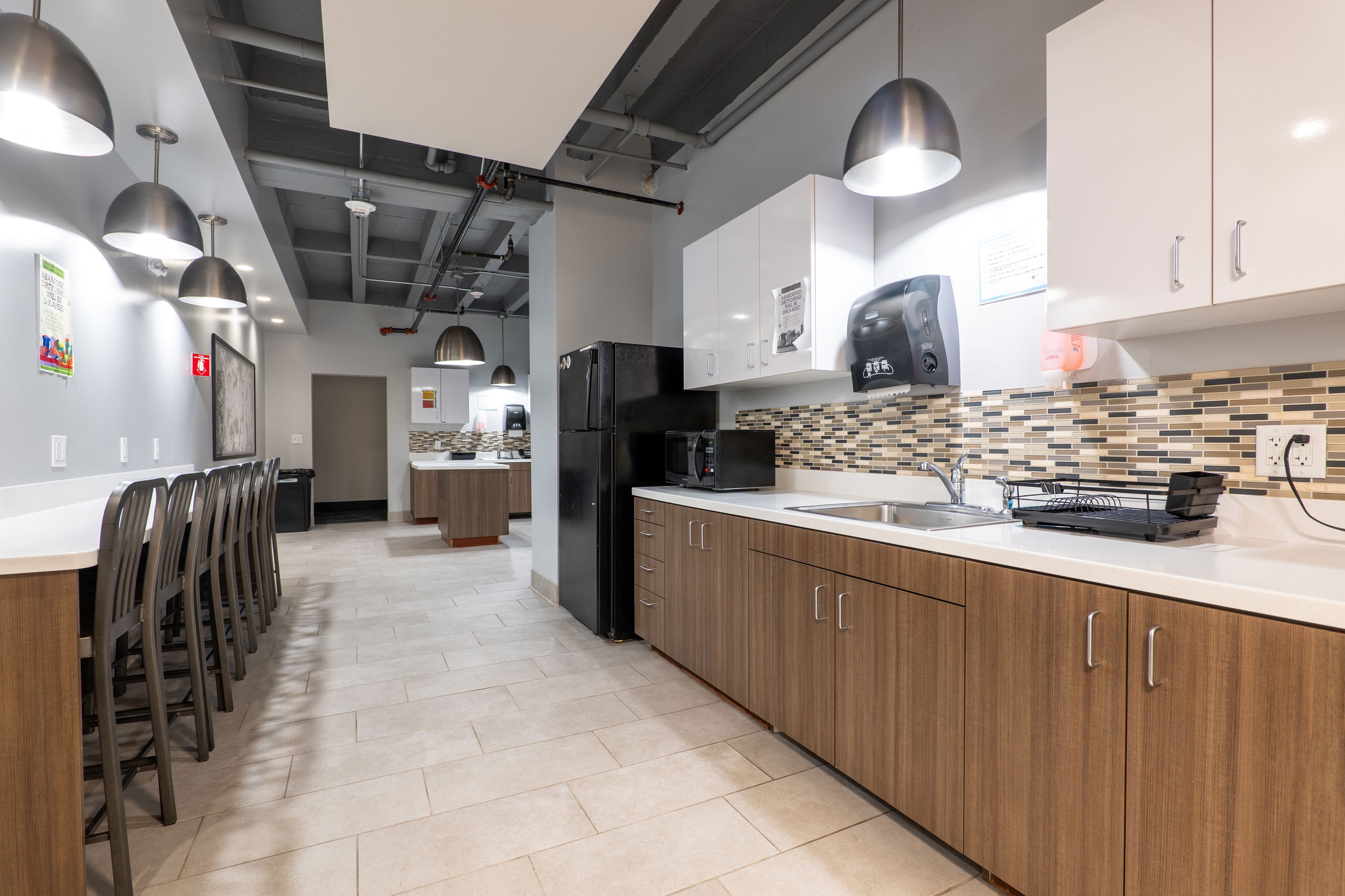 Communal kitchen showing a counter with a sink and microwave, fridge and freezer, and chairs and a counter for personal use