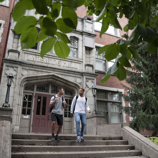 Two Willamette students walk down the steps of the Lausanne Hall dorm, a brick building surrounded by foliage