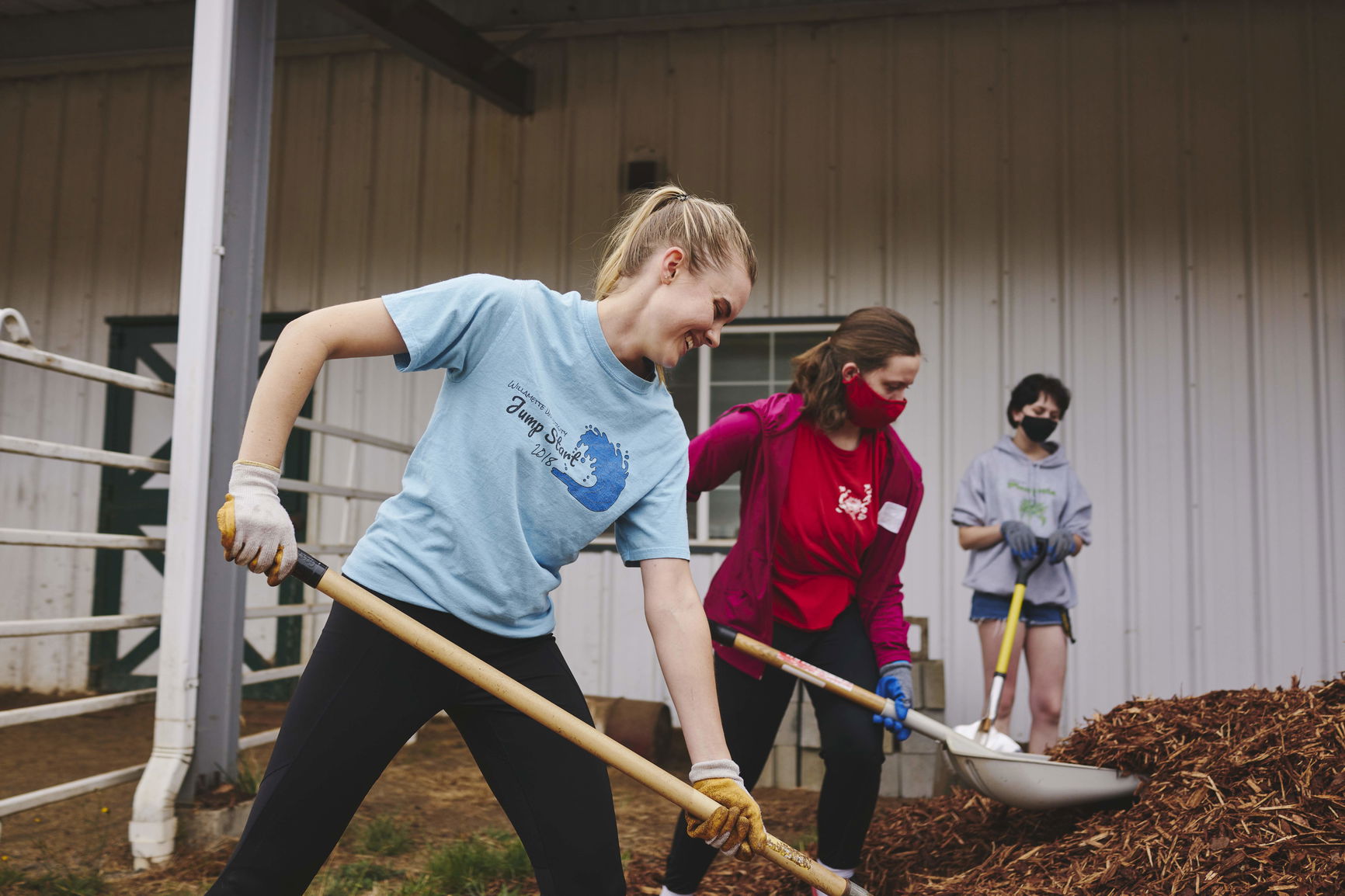 Three Willamette students shovel hay in front of a barn as part of a summer community service program