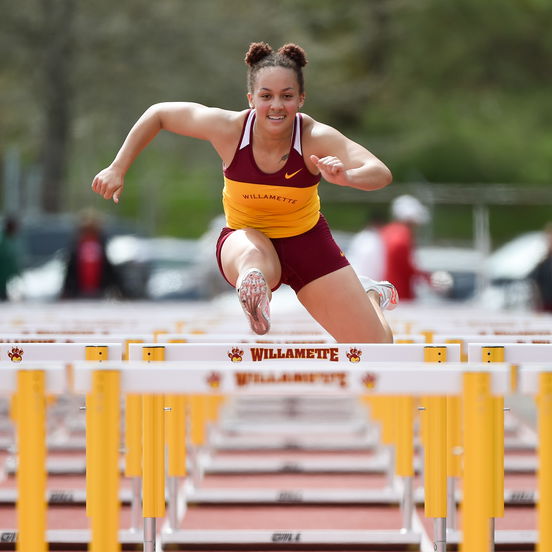 A student athlete in a Willamette uniform leaps over a hurdle on an outdoor track