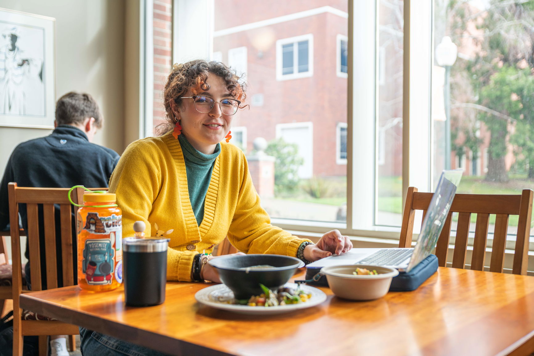 A student in a yellow sweater sits at a table at Willamette's Goudy Commons dining hall with a meal and her laptop