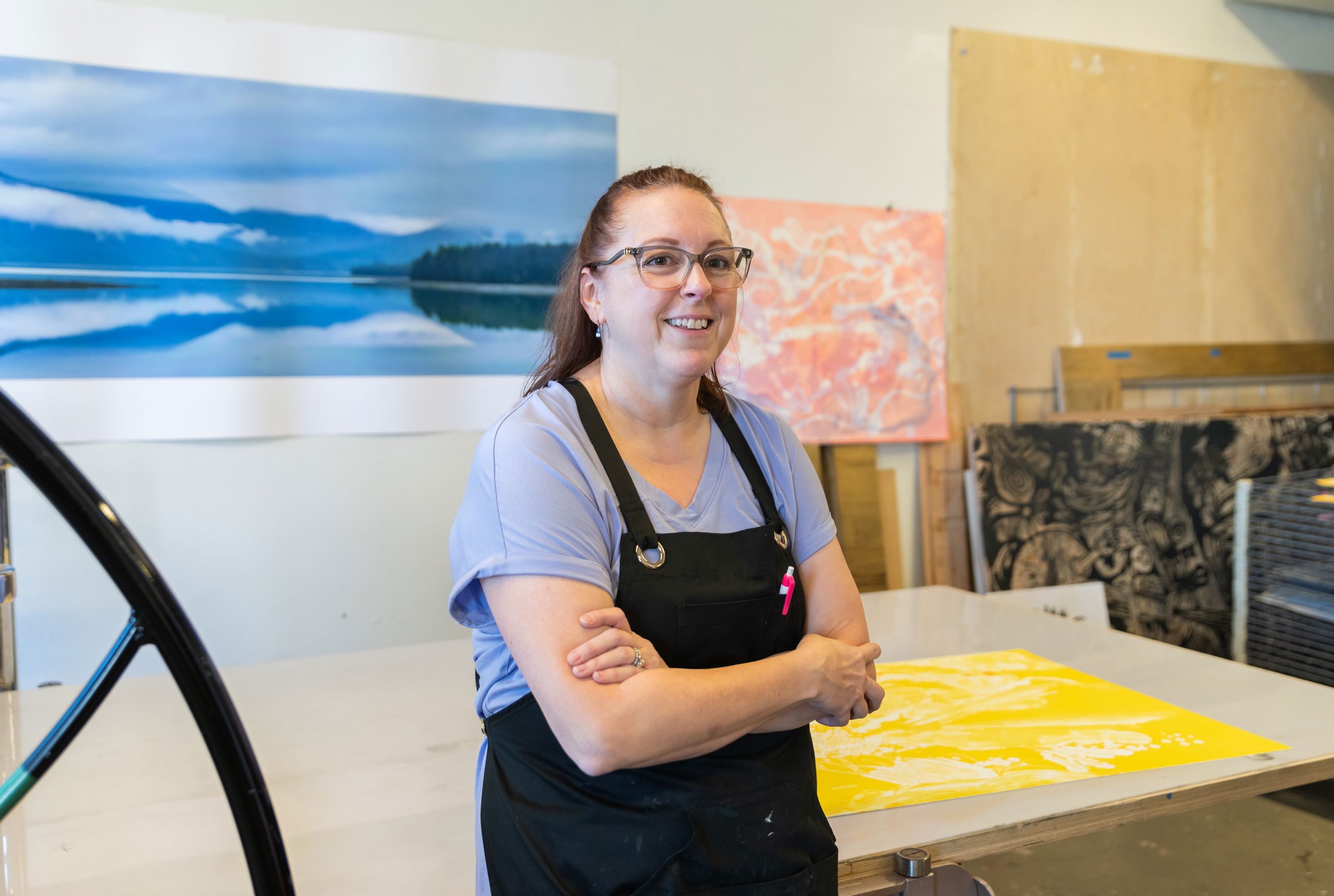 A woman wearing an apron leans against a table in PNCA's printmaking studio