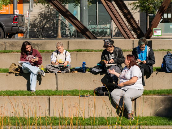 A group of PNCA students sit on grass-covered concrete steps in a park, writing in their notebooks