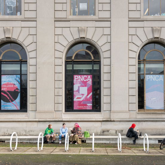 Students sit on a bench in front of PNCA, a white stone building with large arched windows covered with PNCA posters