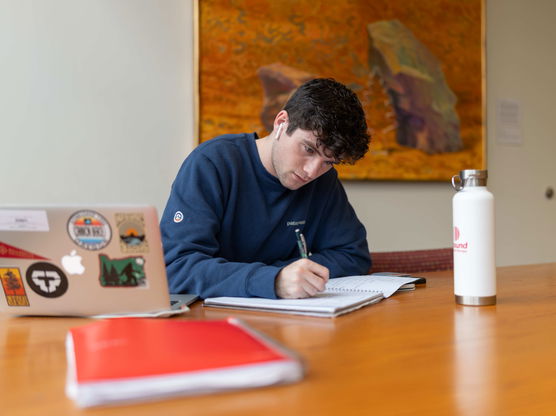 A Willamette student sits at a table in Hatfield Library taking notes in a notebook with his laptop in front of him