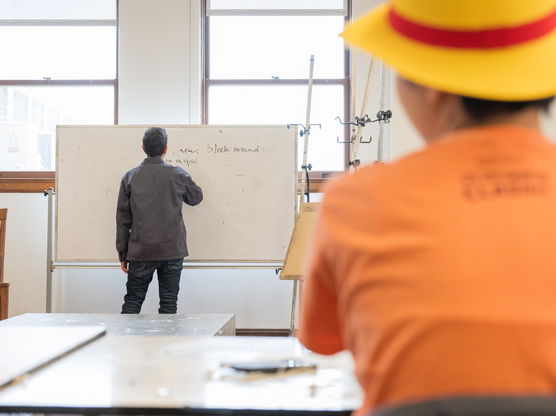 A PNCA professor writes on a whiteboard as a student in a yellow hat watches