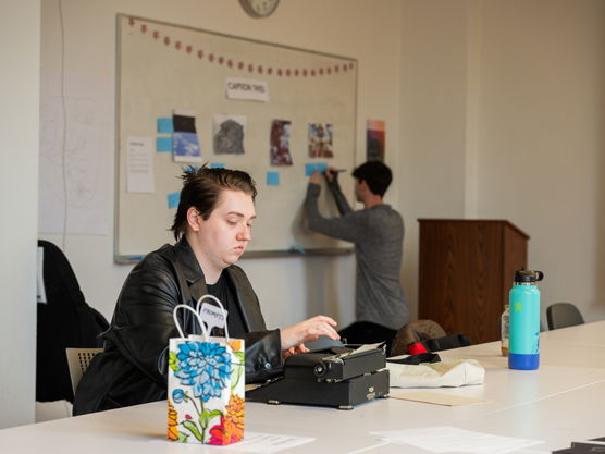 A PNCA student sits at a table in a classroom using a typewriter while another student attaches pages to a bulletin board