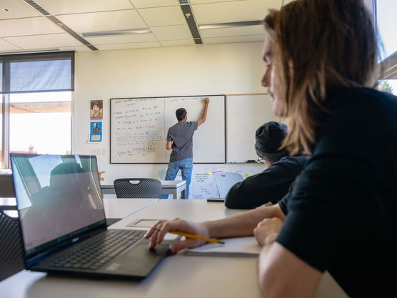 A Willamette student works on a computer while in the background a professor writes on a whiteboard