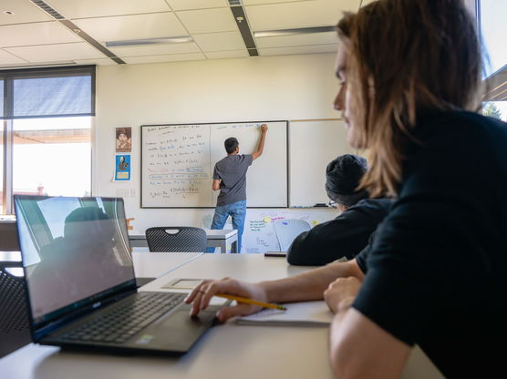 A Willamette student works on a computer while in the background a professor writes on a whiteboard