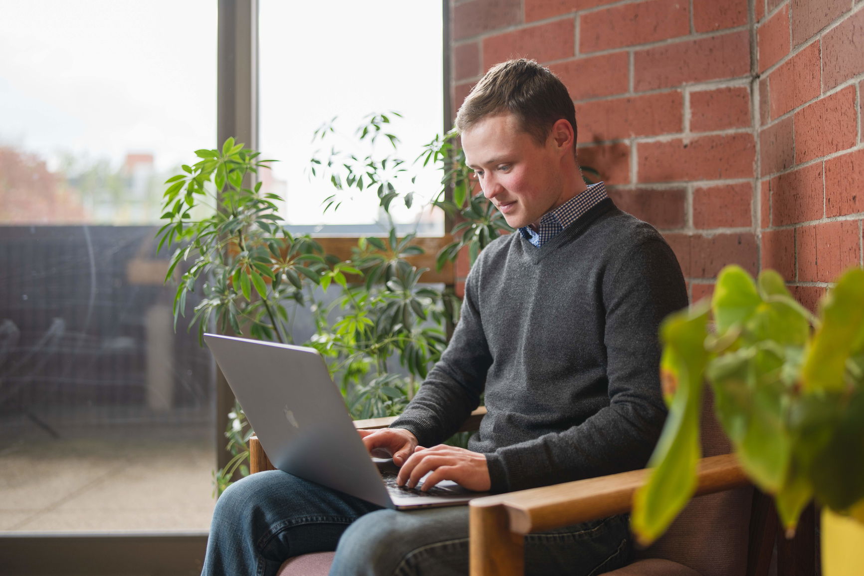 A Willamette MBA student sits in a chair in front of a brick wall and glass sliding door, working on a laptop