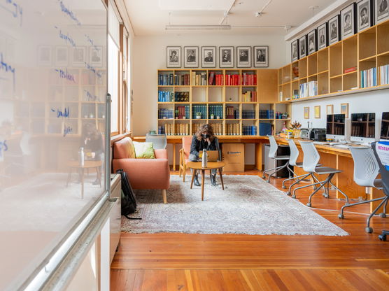 A PNCA student sits at a coffee table and looks at her laptop in a classroom with computer desks, bookshelves, and whiteboard