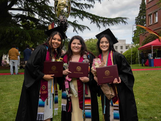 Three Willamette undergrad students wearing graduation robes at commencement pose outside on campus holding their diplomas