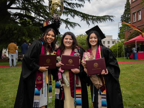 Three Willamette undergrad students wearing graduation robes at commencement pose outside on campus holding their diplomas