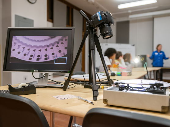 A camera on a tripod and a computer monitor sit on a table in the back of a classroom where a PNCA professor is teaching