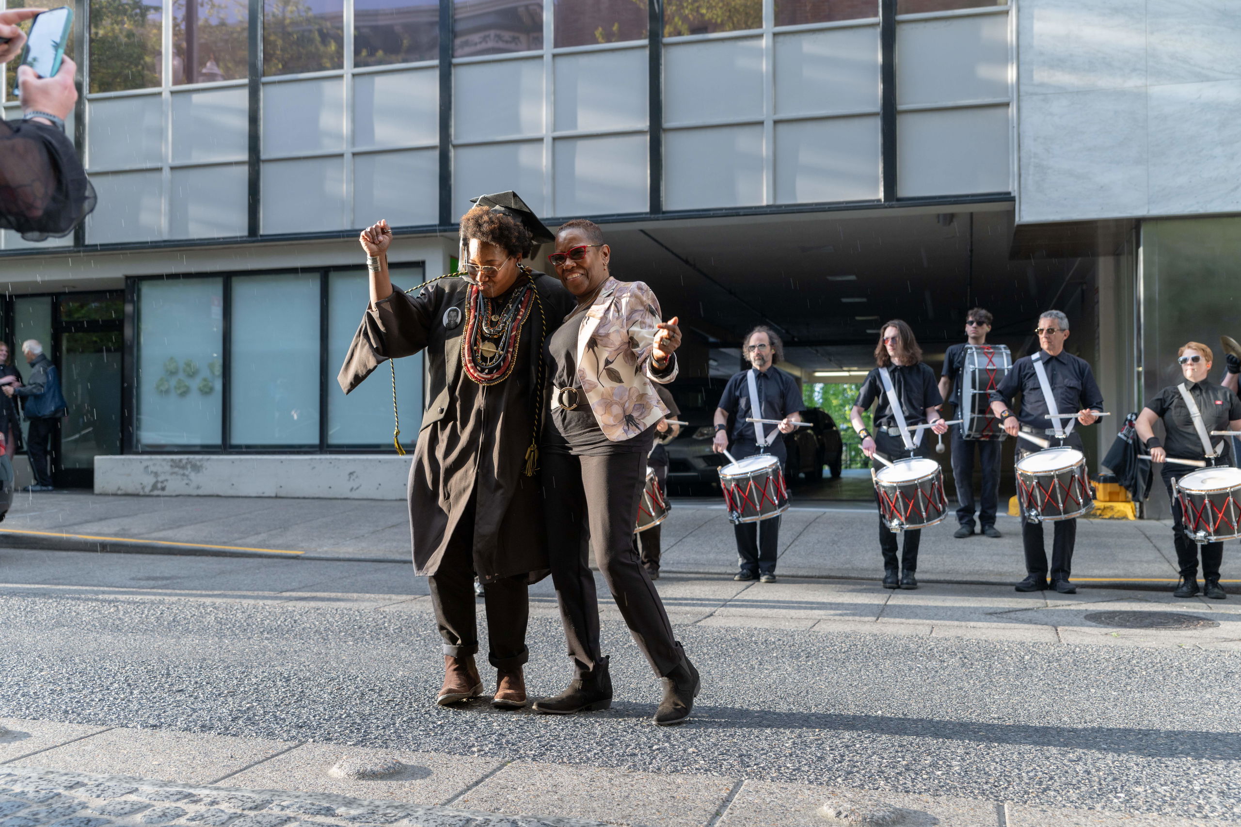 At PNCA's commencement, a student in a graduate robe dances on the street with a family member in front of a drum line