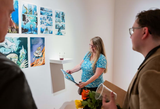 Three people examine prints of a comic book displayed on a wall of a PNCA art gallery