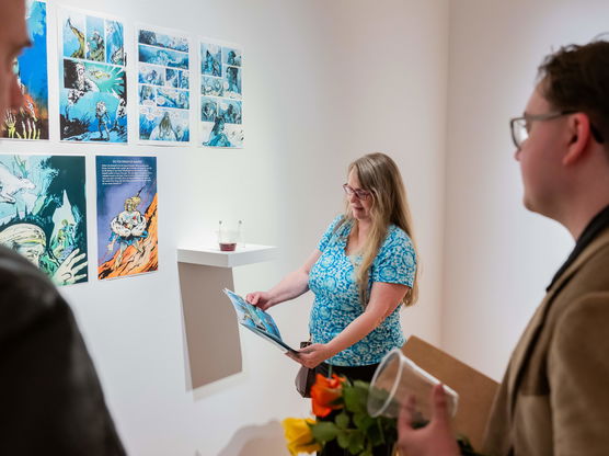 Three people examine prints of a comic book displayed on a wall of a PNCA art gallery