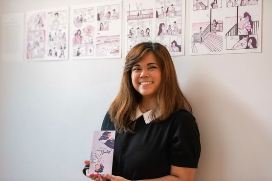 A smiling PNCA student stands in front of four prints of graphic novel pages and holds a card with a similar illustration