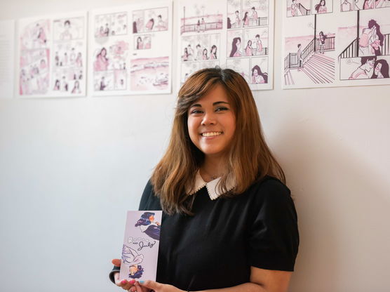 A smiling PNCA student stands in front of four prints of graphic novel pages and holds a card with a similar illustration