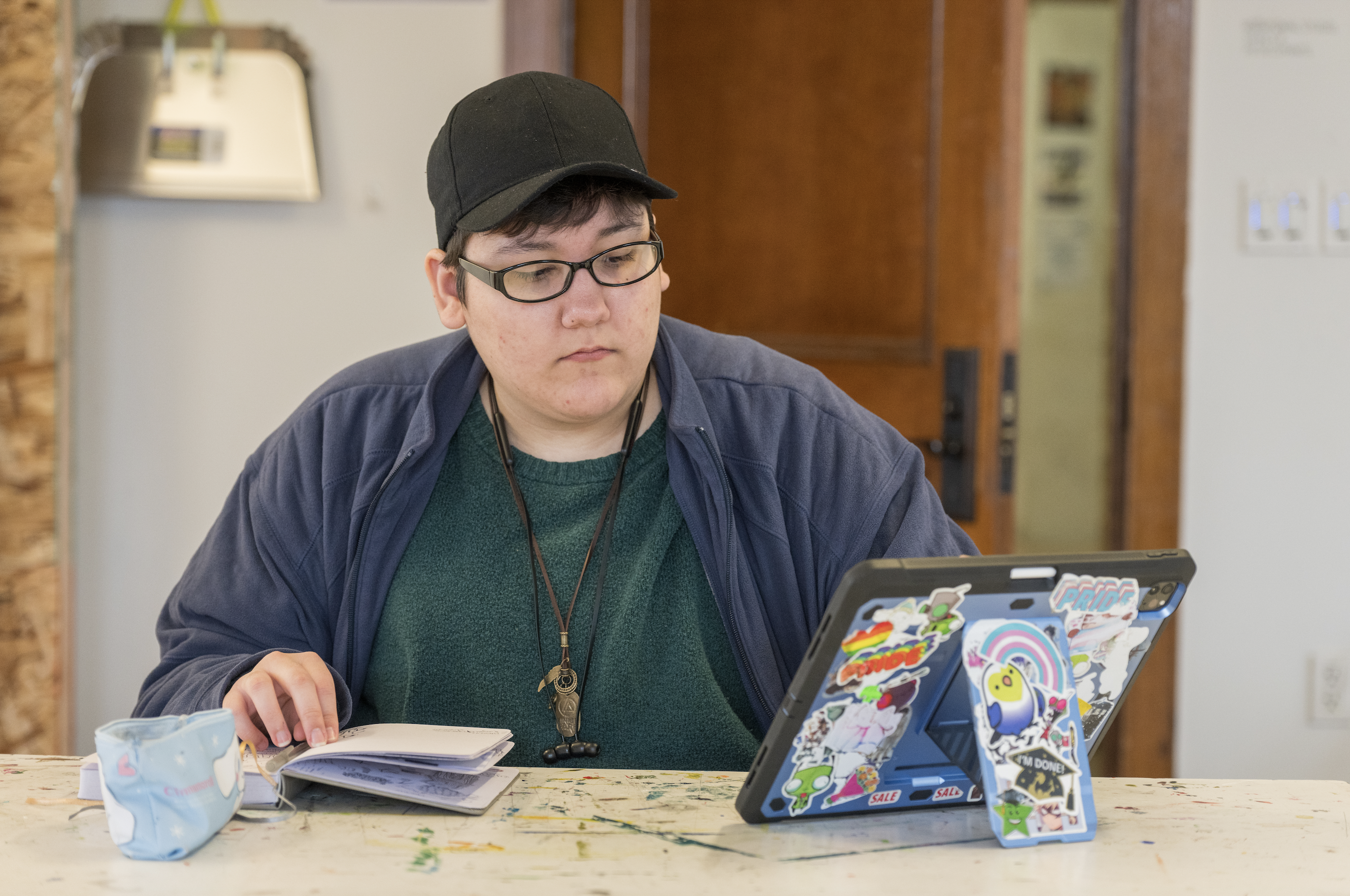 A PNCA student sits at a counter and looks at a tablet covered in stickers while taking notes in a notebook