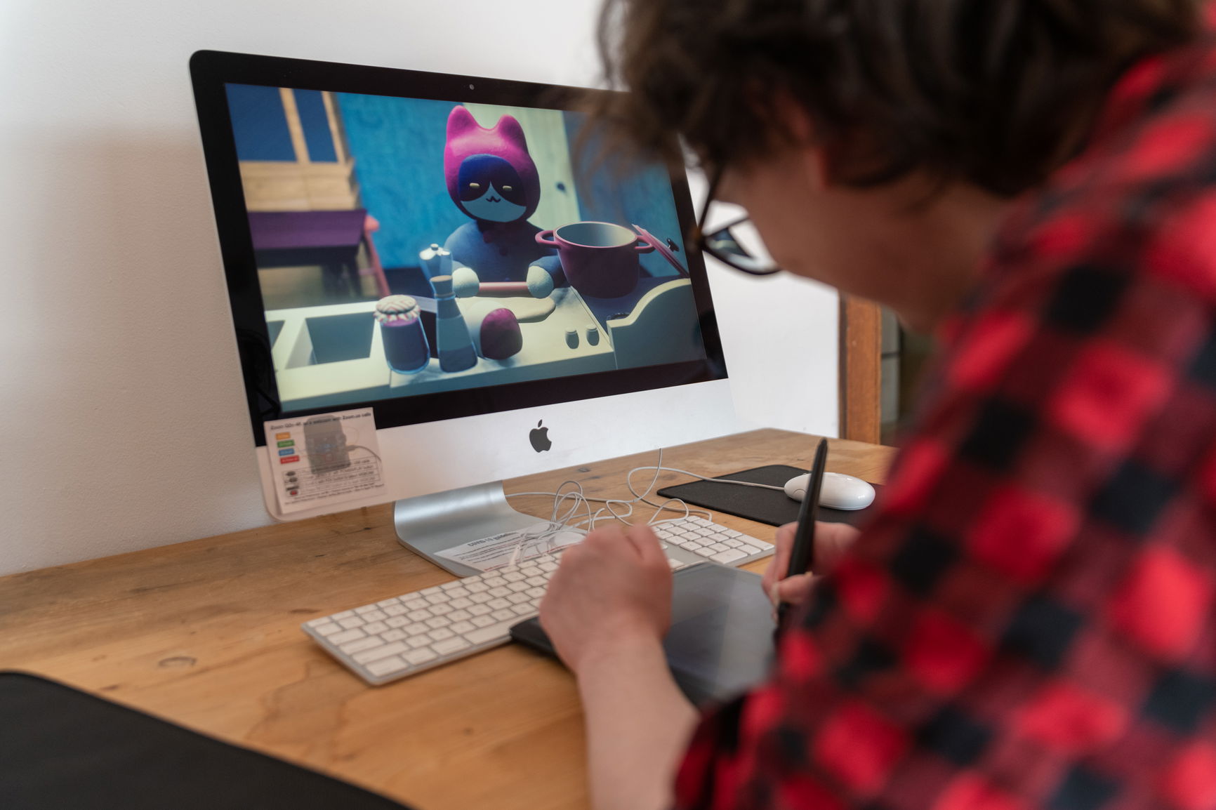 A person digitally drawing in front of a Mac computer with an animated cat in a red hat that is cooking