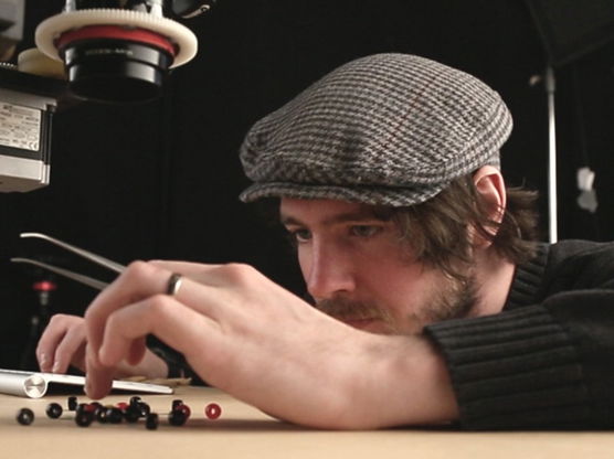 Man wearing a grey flat cap and black sweater, using tweezers to arrange small beads on a table with a camera mounted above.