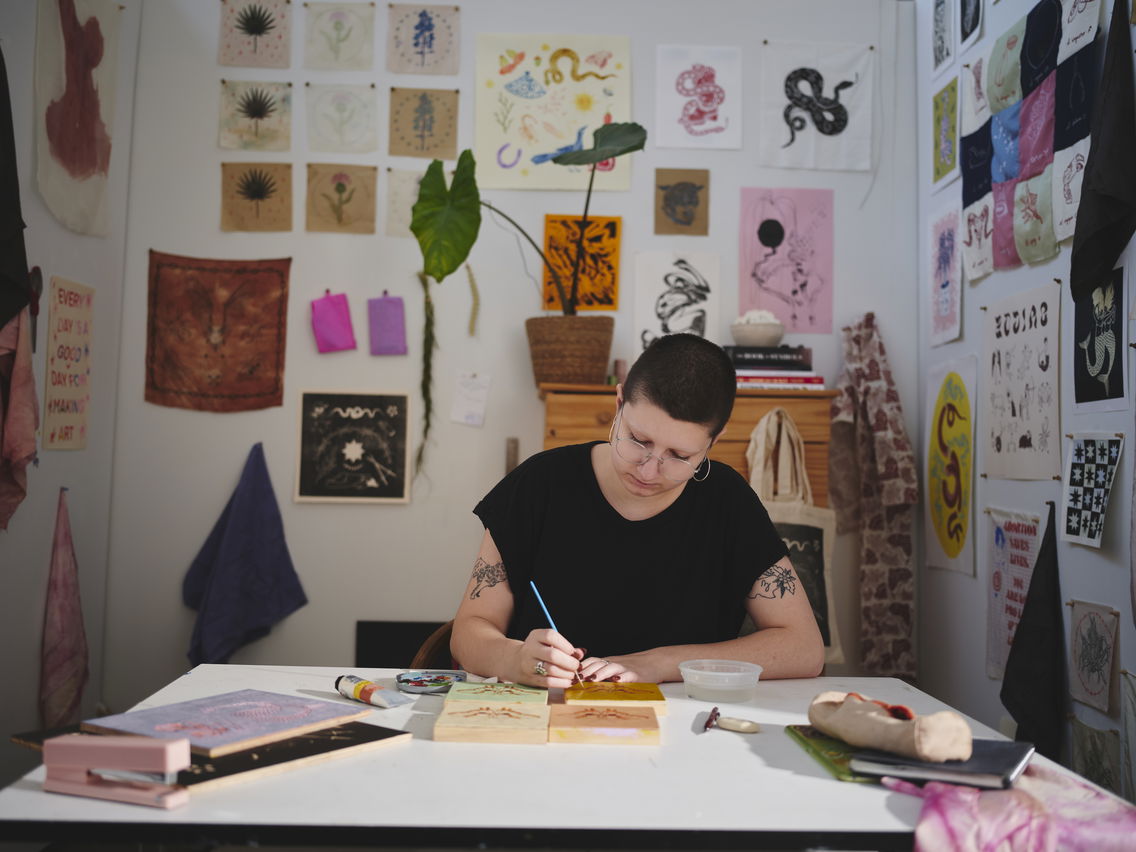 Artist painting at a cluttered desk in a studio with art-covered walls.