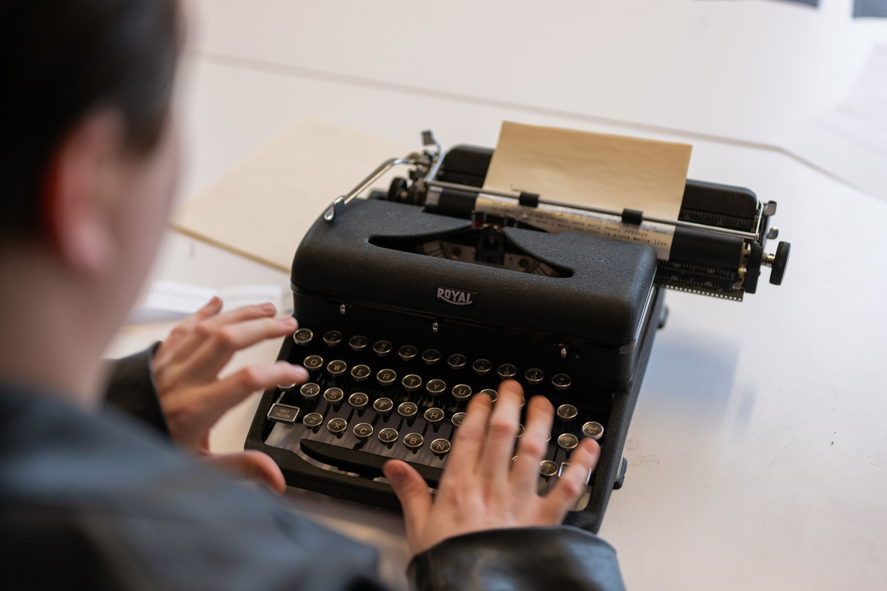 Two people working on a typewriter; one points and explains while the other looks on. Another person works in the background.