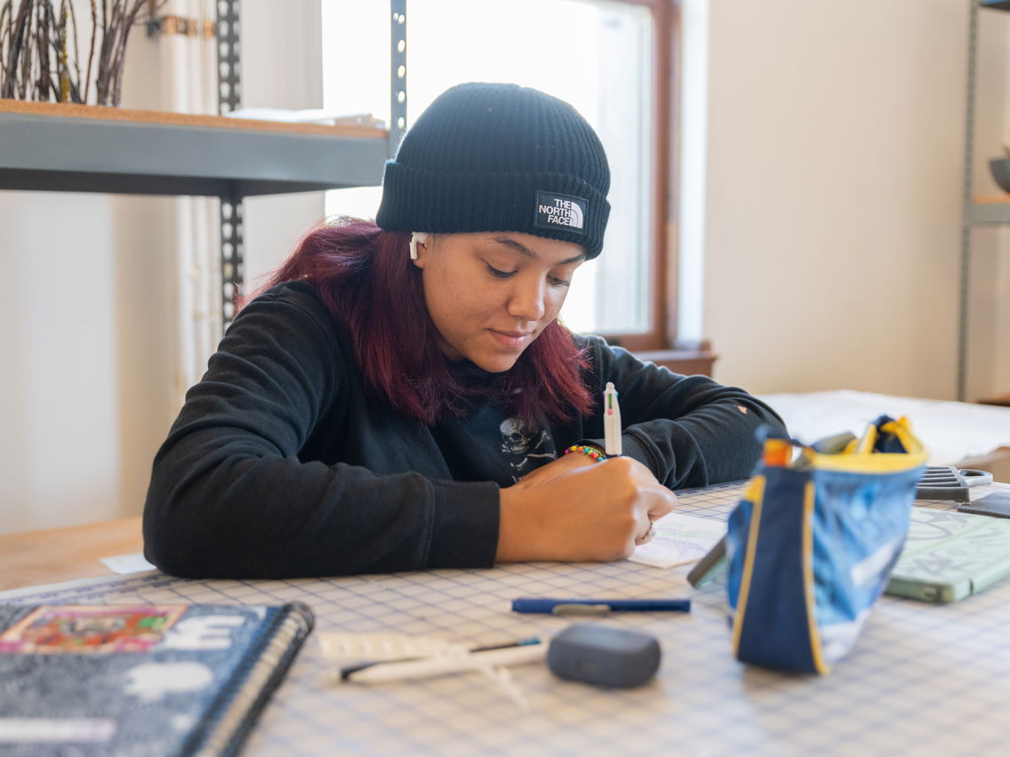 Person wearing a black beanie writing at a table in a well-lit room.