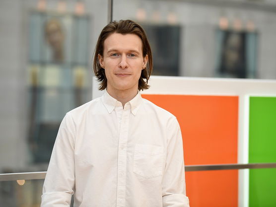 Young man in a white button-up shirt standing indoors with colorful rectangles and blurred windows in the background.