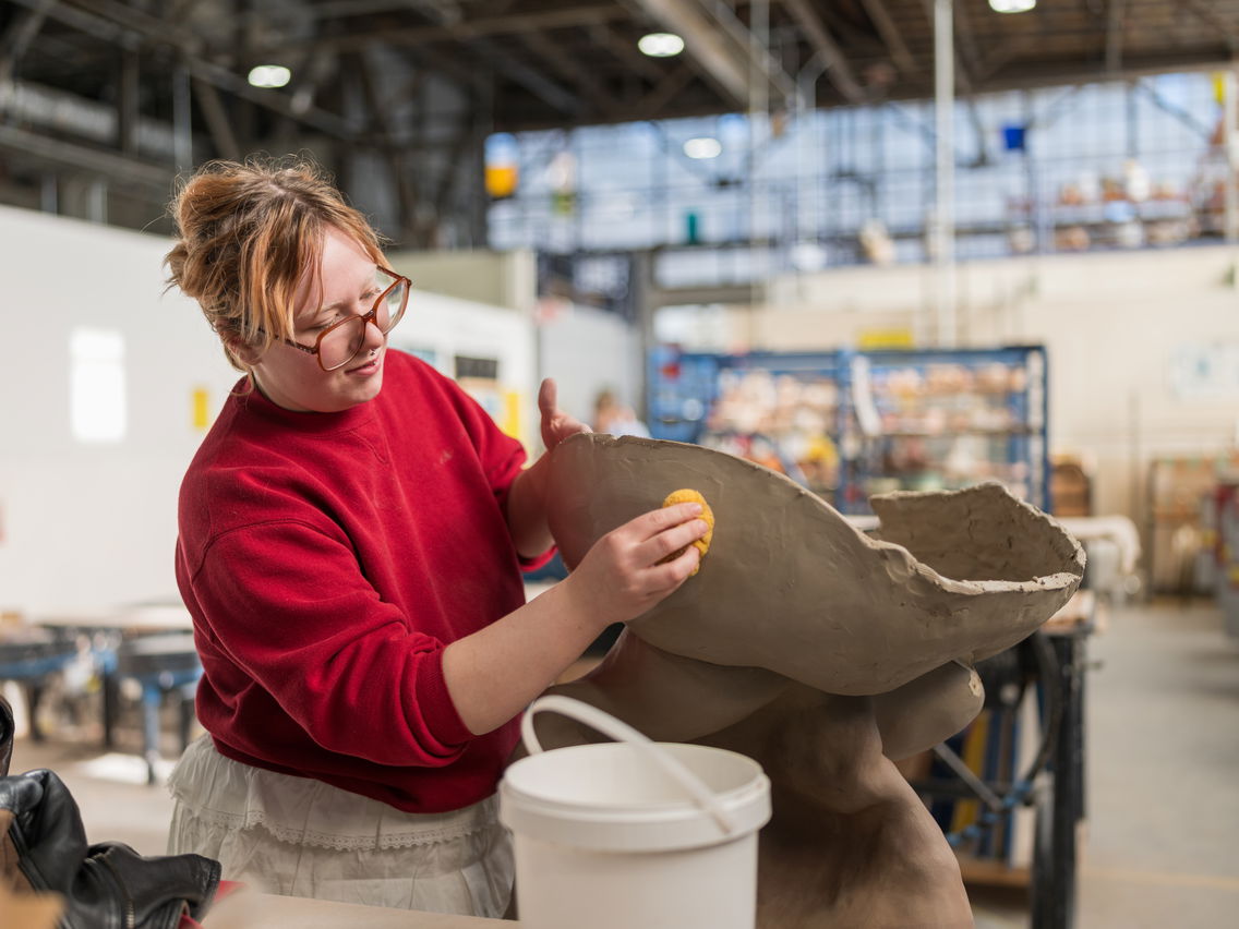 Person smoothing a large clay sculpture with a yellow sponge in a workshop.