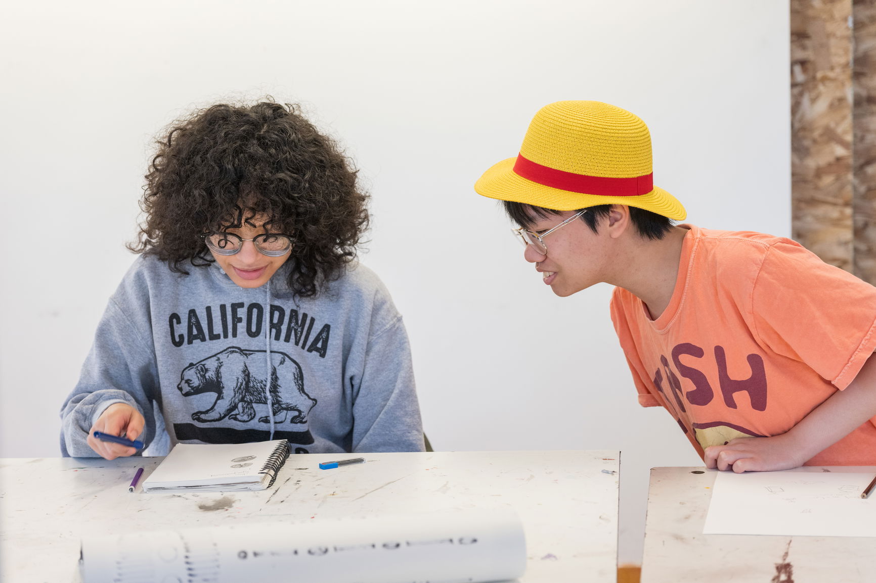 One student with curly hair and another student in a yellow hat looking at a notebook 