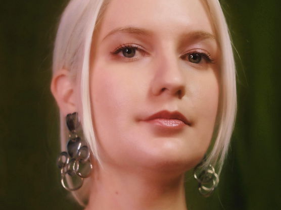 Portrait of a person with platinum blonde hair, wearing large circular metallic earrings, and a black top.